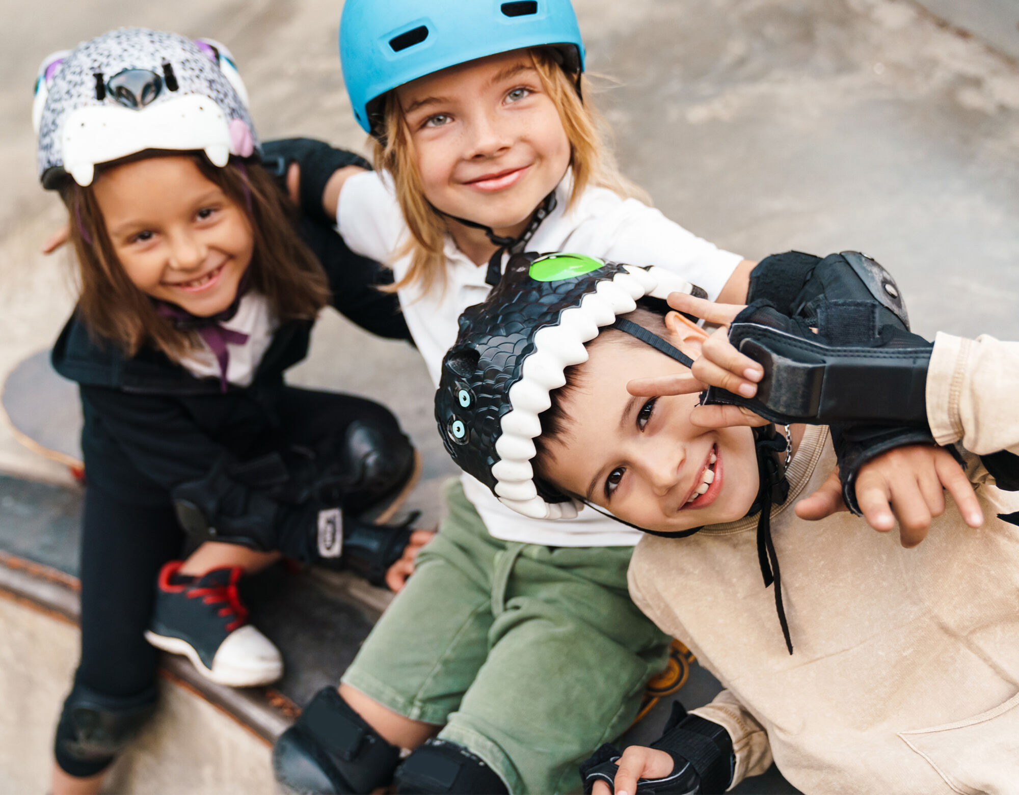 Three children wearing kneepads and helmets smile while sitting on skateboards.