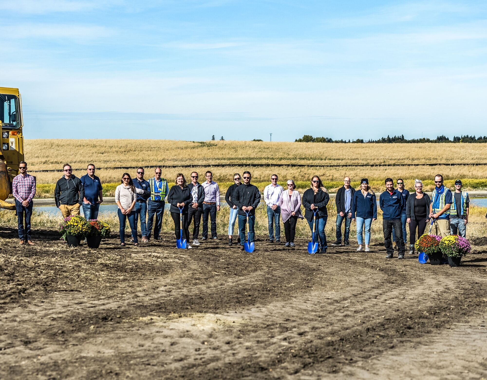 A large group of people stand in an empty field, some of which hold blue shovels.