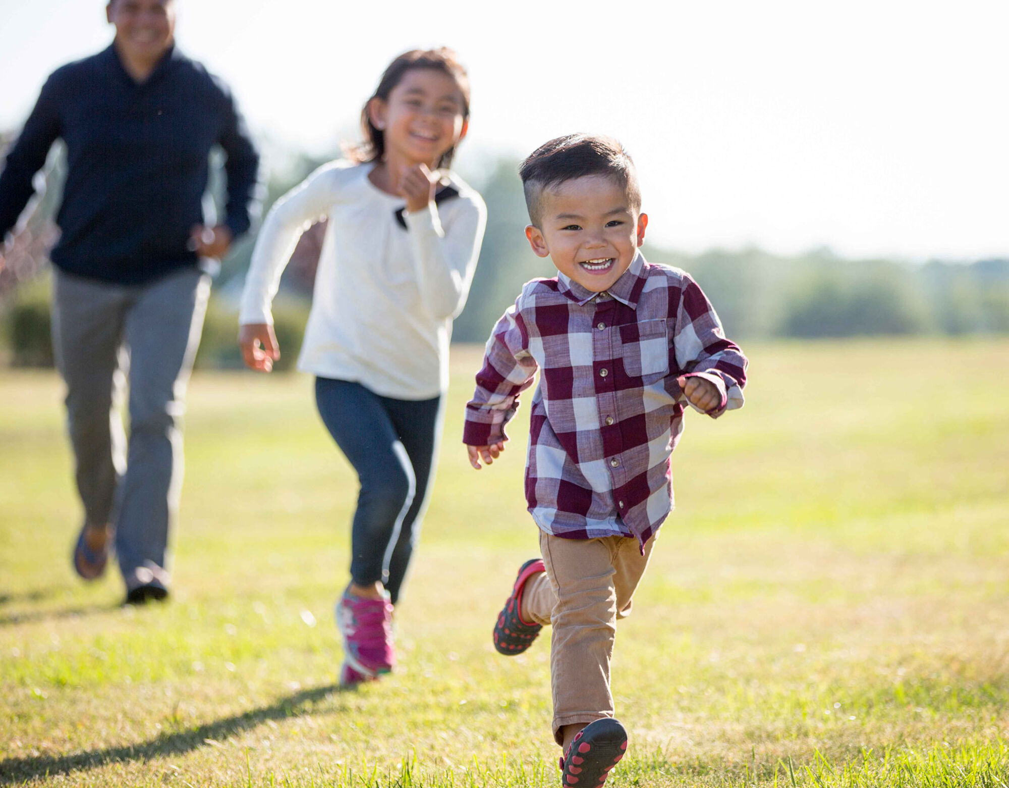 Two children running and smiling on grass with a man running behind them.