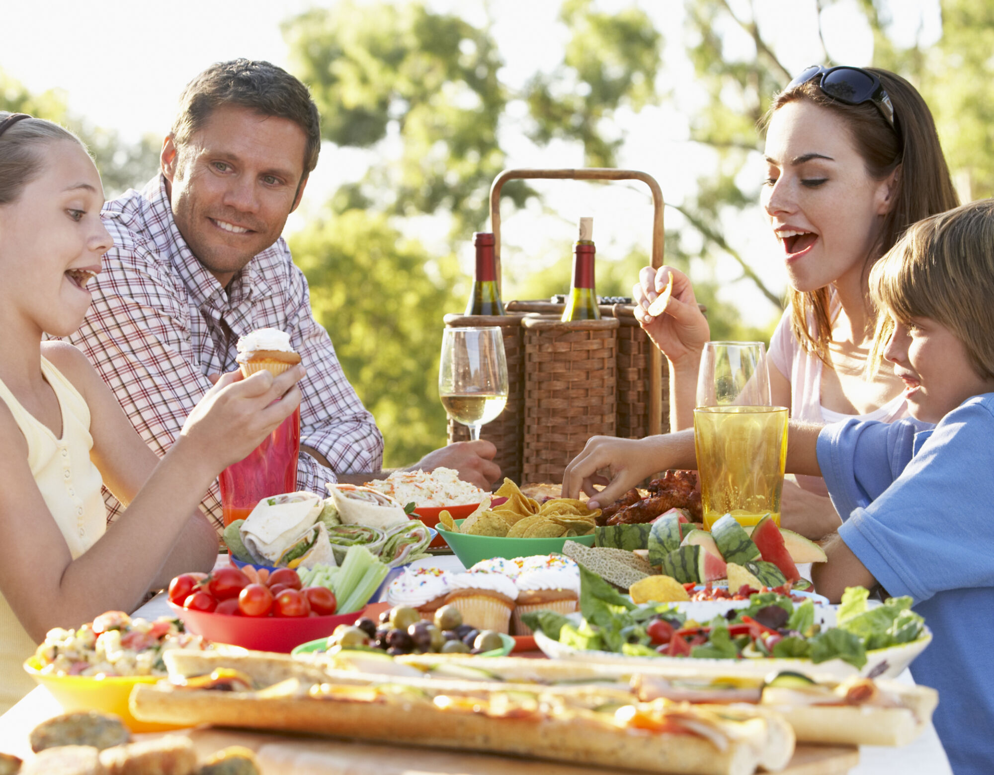 A family shares a large meal on a picnic table outdoors on a sunny day.