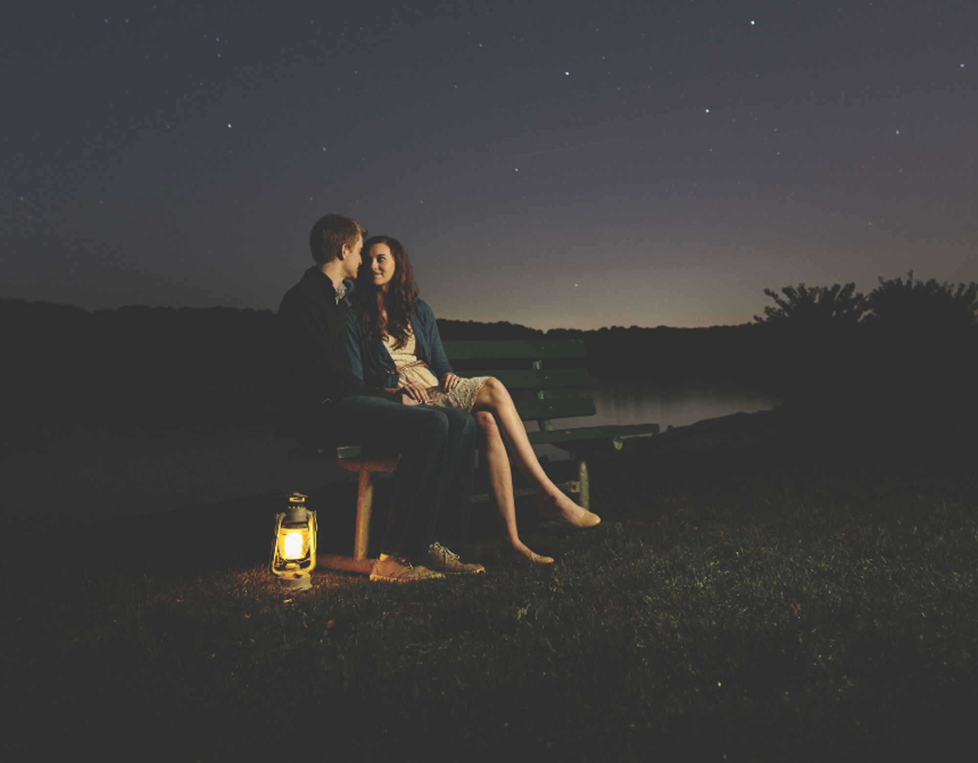 A couple sits on a bench near a lake at night with a bright lantern at their feet.