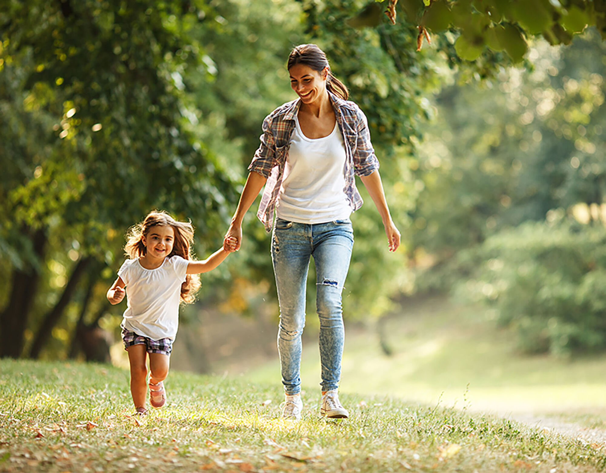 A mother and daughter running through a grassy area surrounded by trees.