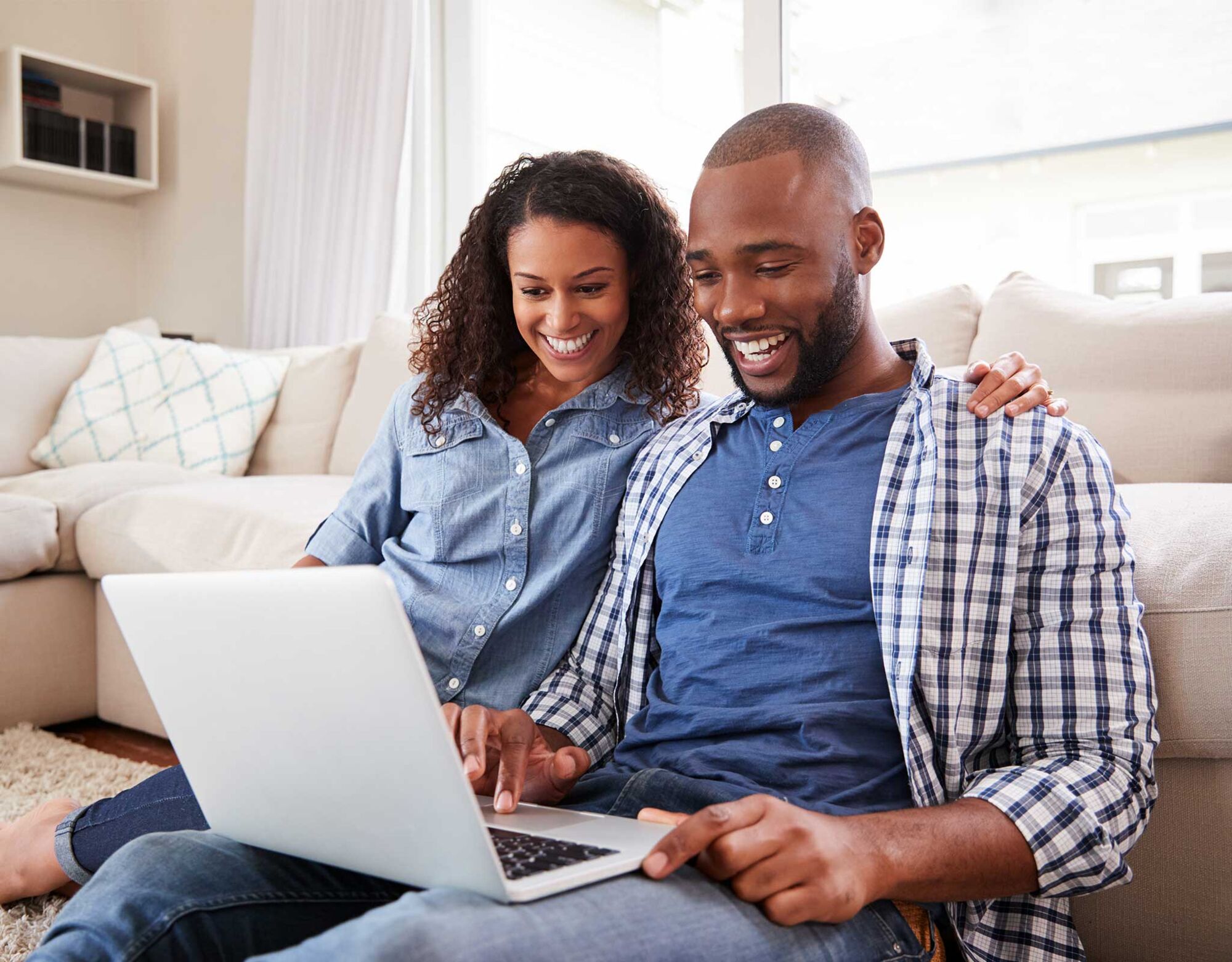 A couple sitting on the floor looking at a laptop and smiling.