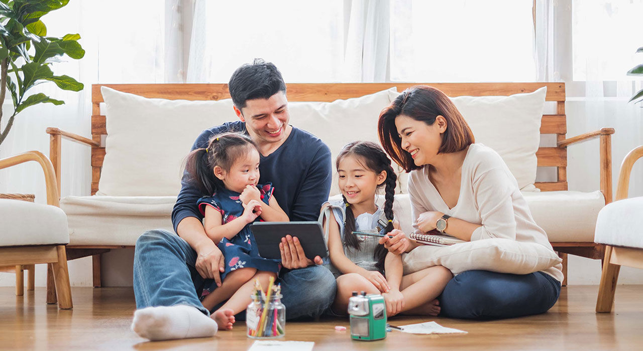 A family playing on a tablet with their two children on the wooden floor.