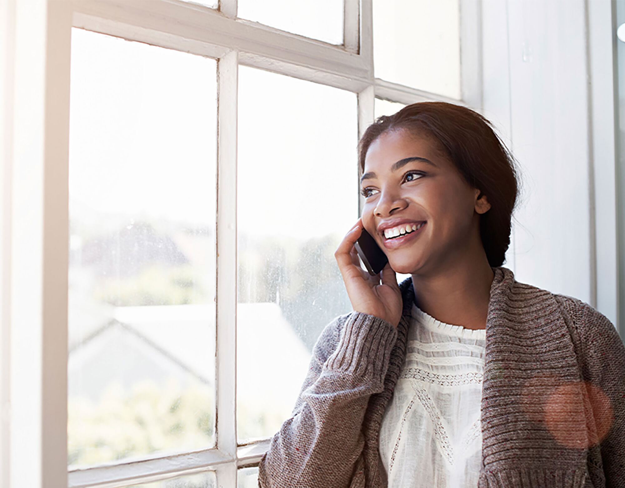 A woman beside a window on her cell phone and smiling. 