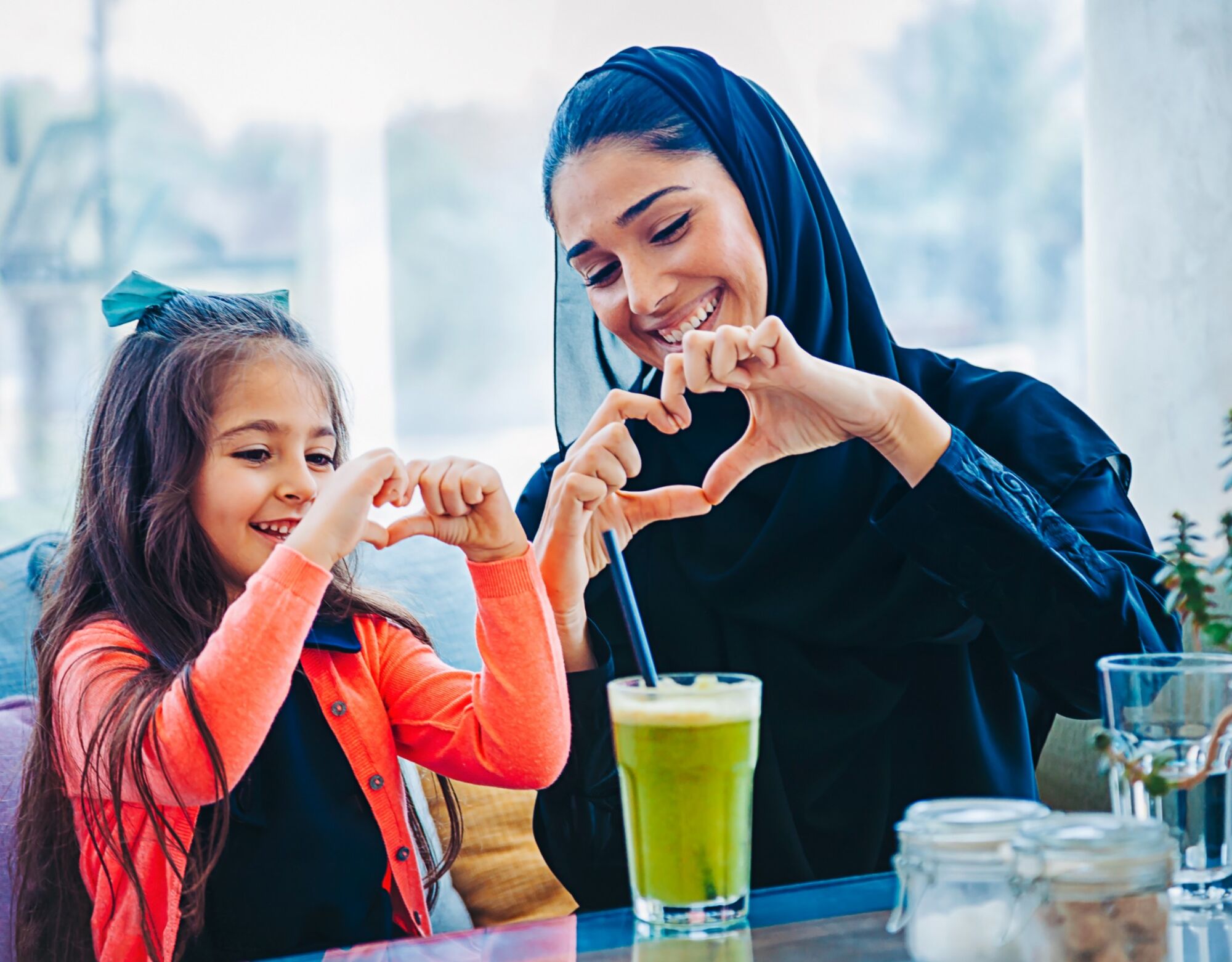 A mother and young daughter make hearts with their hands while a green juice is on the table in front of them.