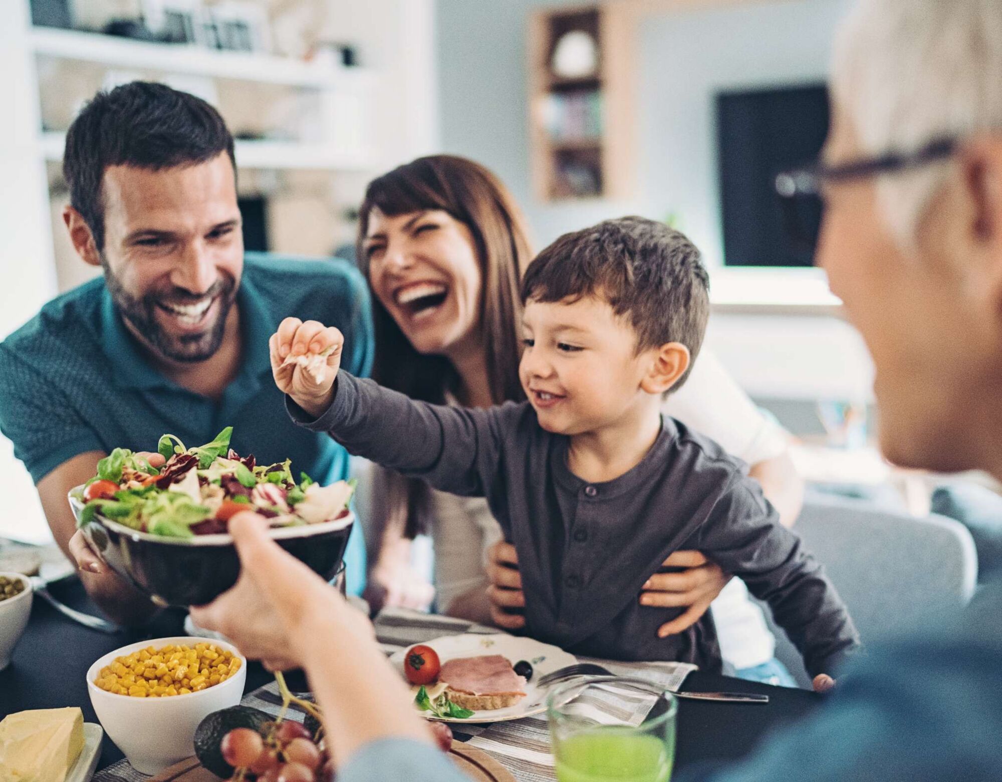 A toddler takes a leaf out of a salad bowl at the dining table surrounded by his parents and grandparents.