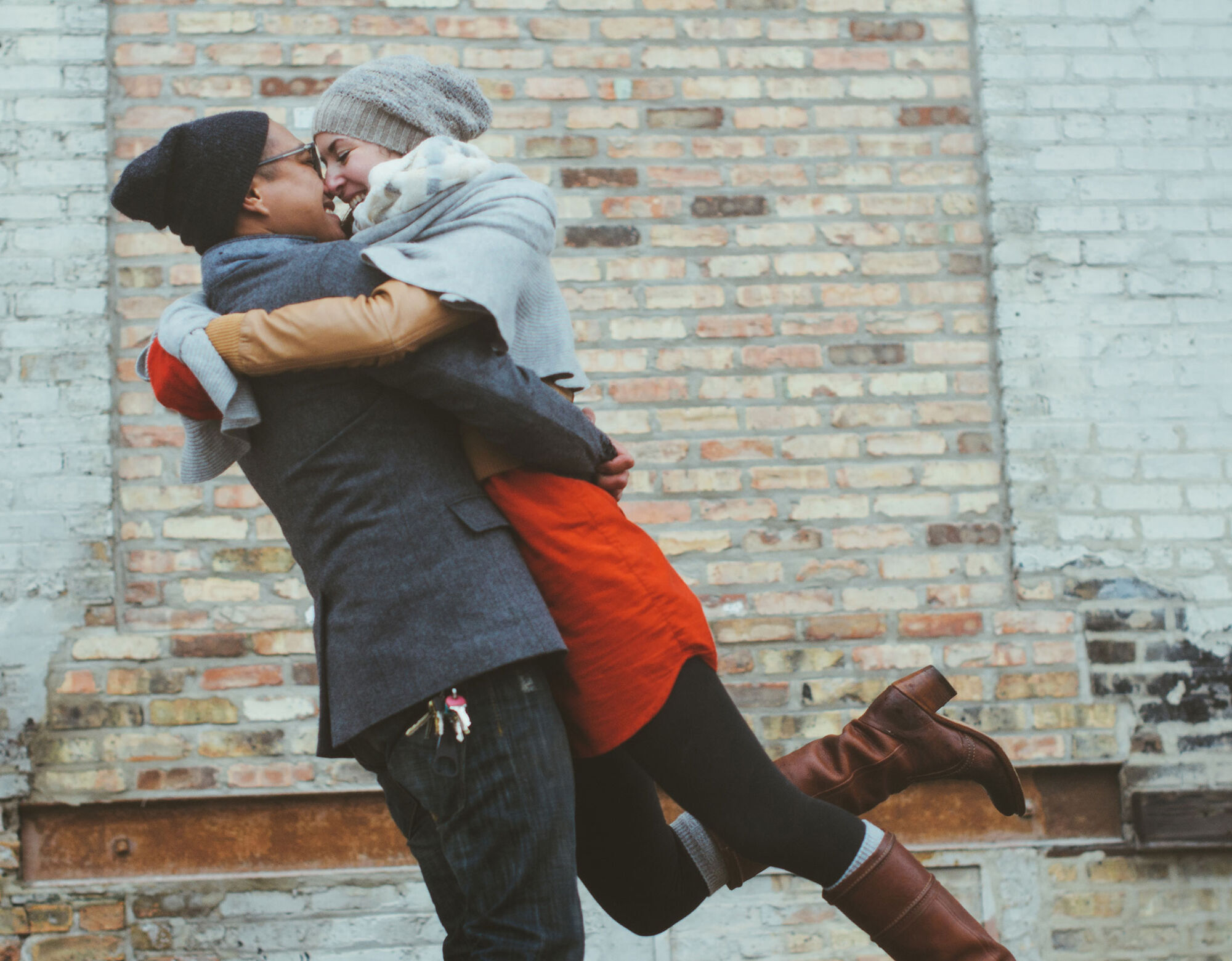 A man holds a woman up happily as they embrace in front of a brick wall.