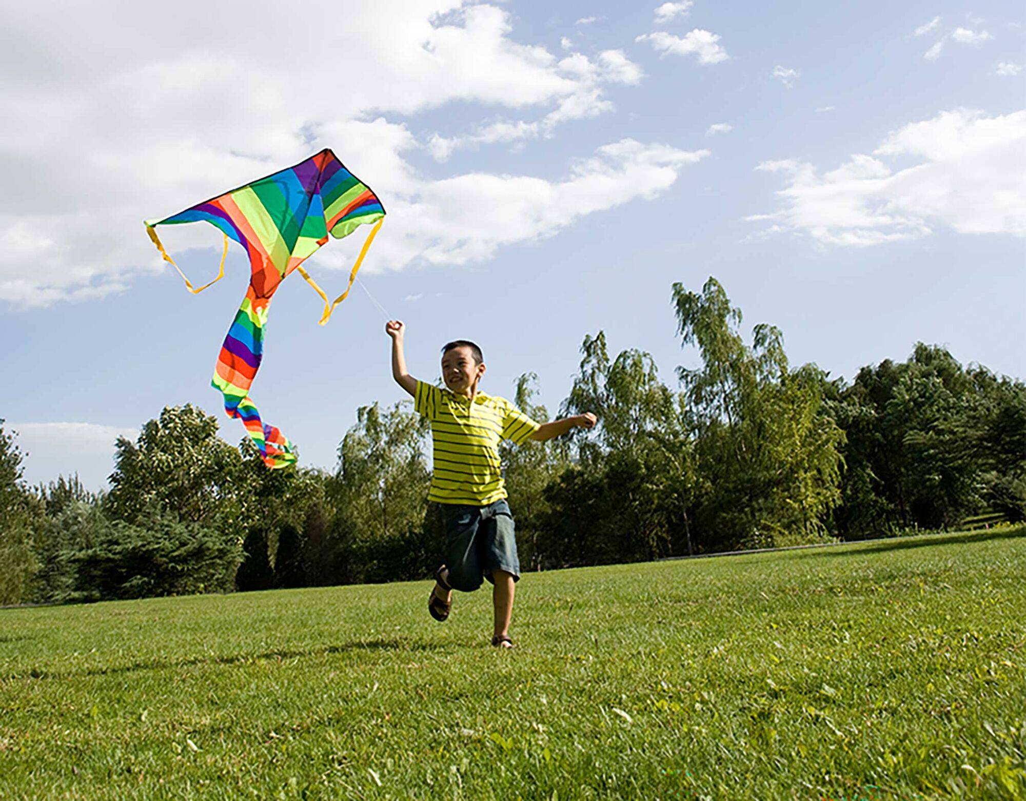 A young boy runs through a field of grass, holding up a large rainbow kite.