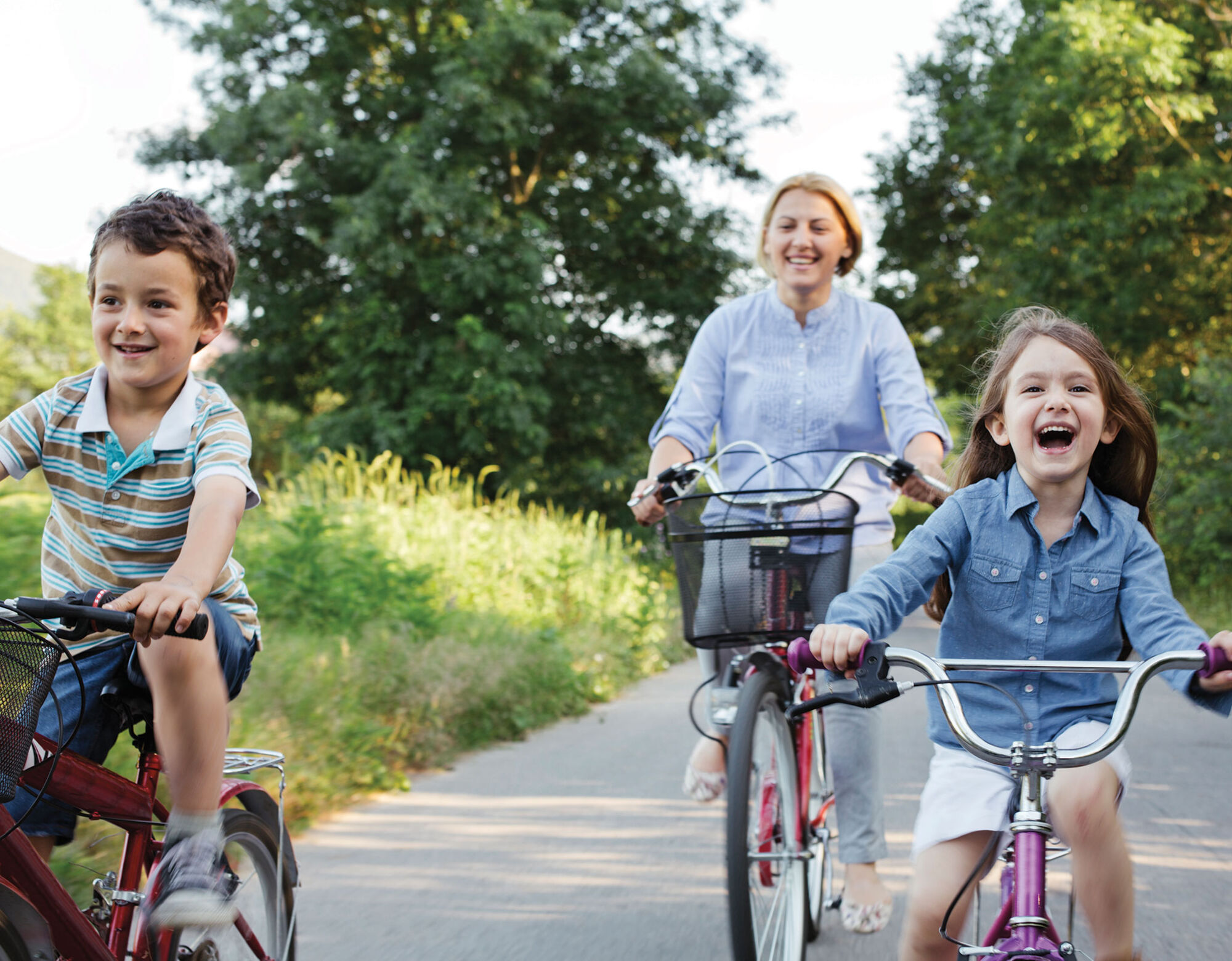 Two children ride bikes down a path surrounded by grass and trees while their mother follows behind on a bike.