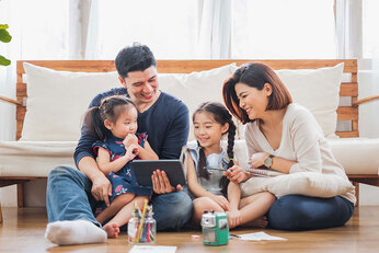 A family playing on a tablet with their two children on the wooden floor.