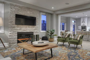 Greatroom with TV and fireplace attached to a brick wall, two chairs off to the right and the kitchen behind. 