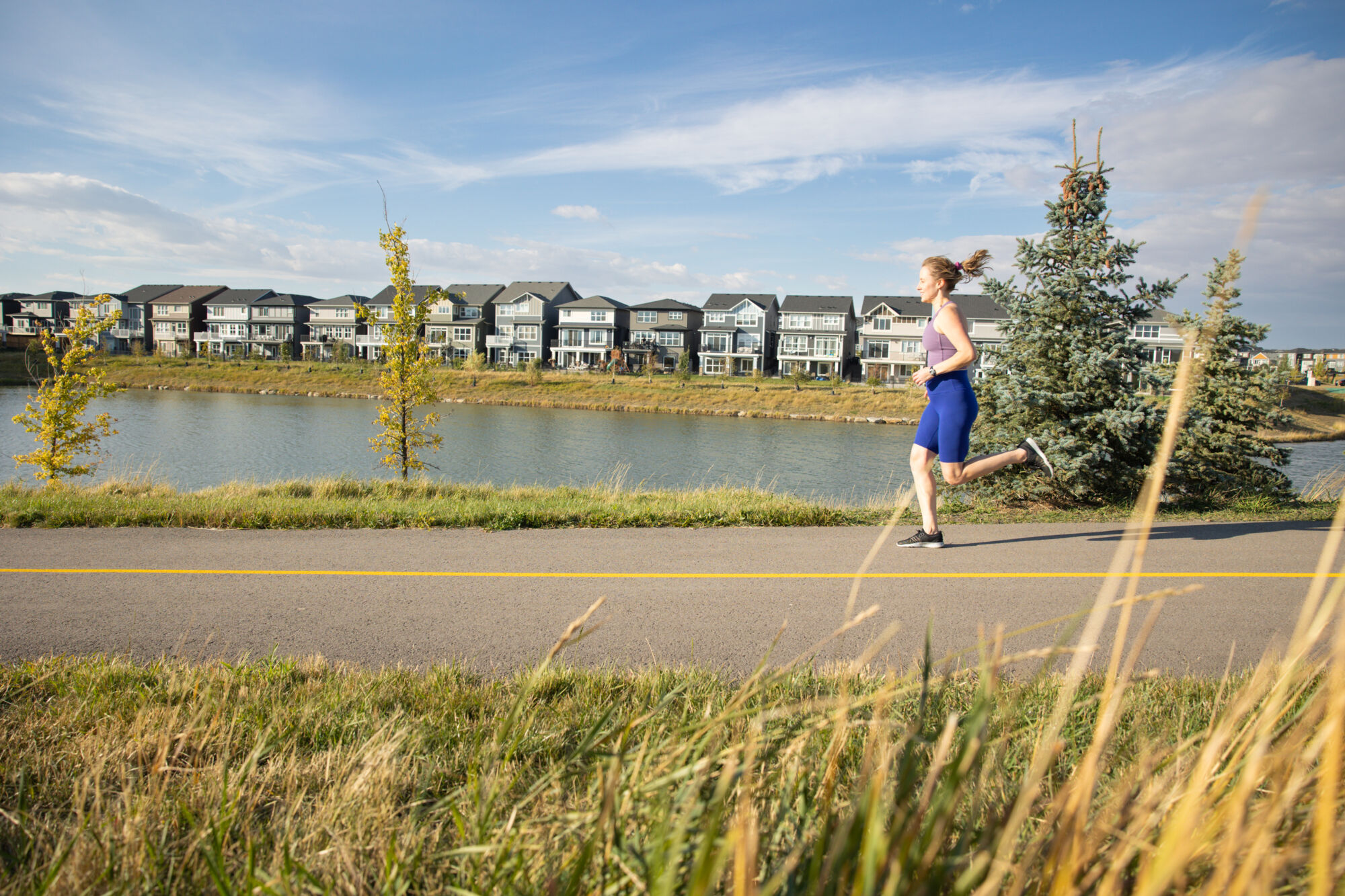 A woman jogging along side some trees along a pathway with a pond and a row of houses in the distance