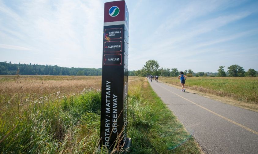 Outdoor path with people running and biking on it, surrounded by grass and trees, with a large directional sign for the Rotary/Mattamy Greenway.