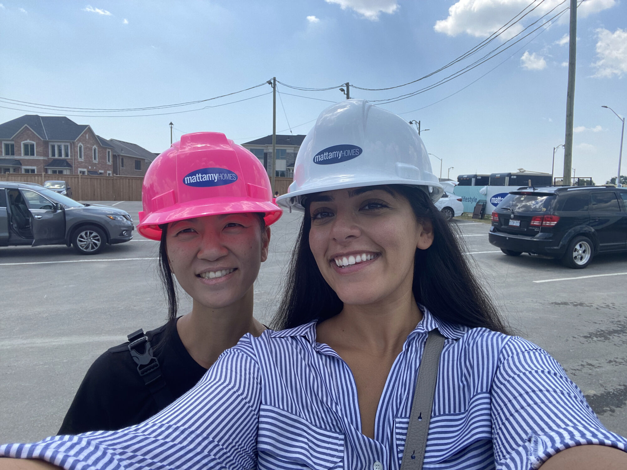Two woman taking a selfie, each wearing a pink and white hard hat with the Mattamy logo on it. With parked cars and houses in the background.