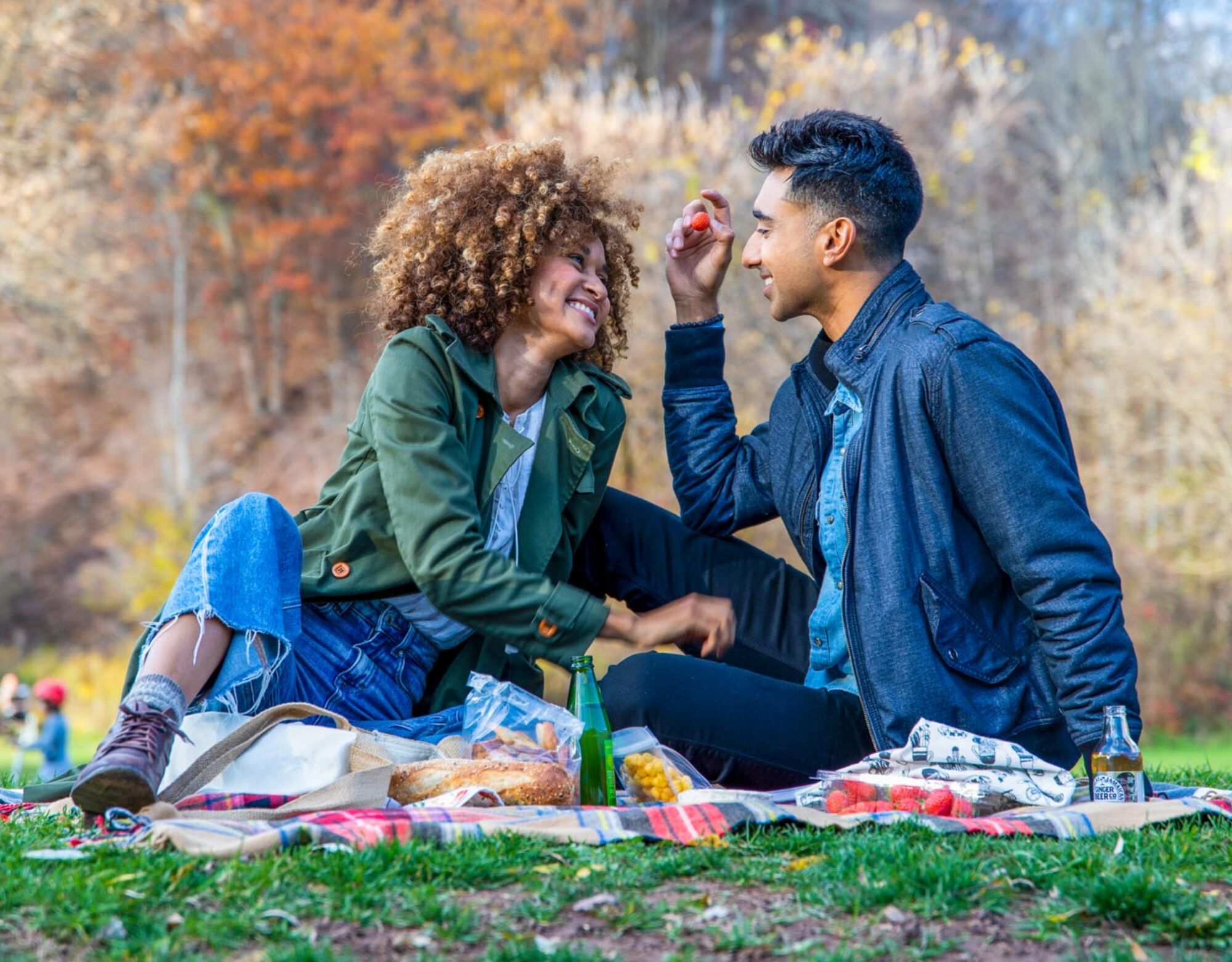 A couple having an outdoor picnic, cuddling on a plaid blanket, with sandwiches, snacks and drinks in front of them.