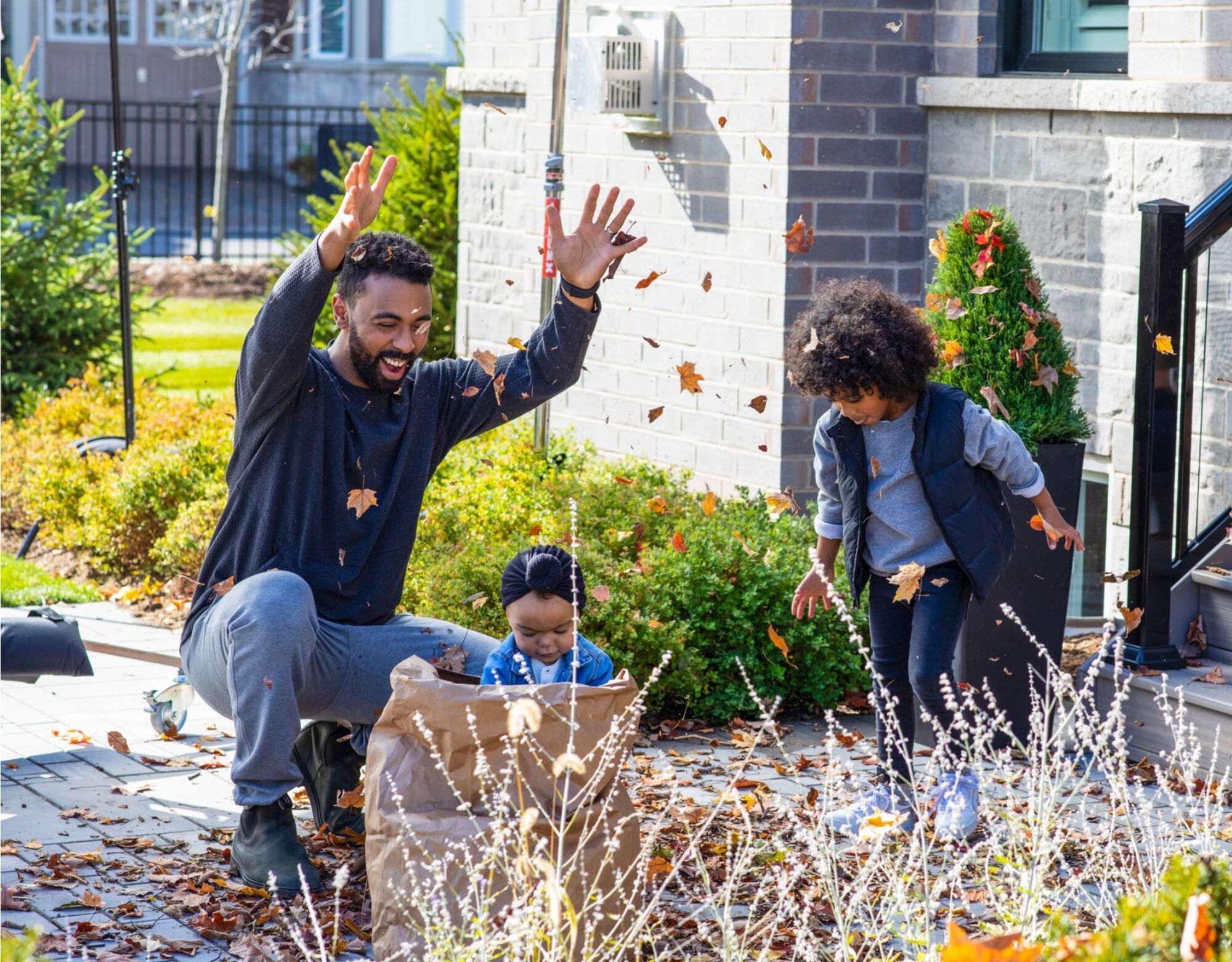 A father and his two children playing in the front yard, throwing leaves up in the air during the fall season.