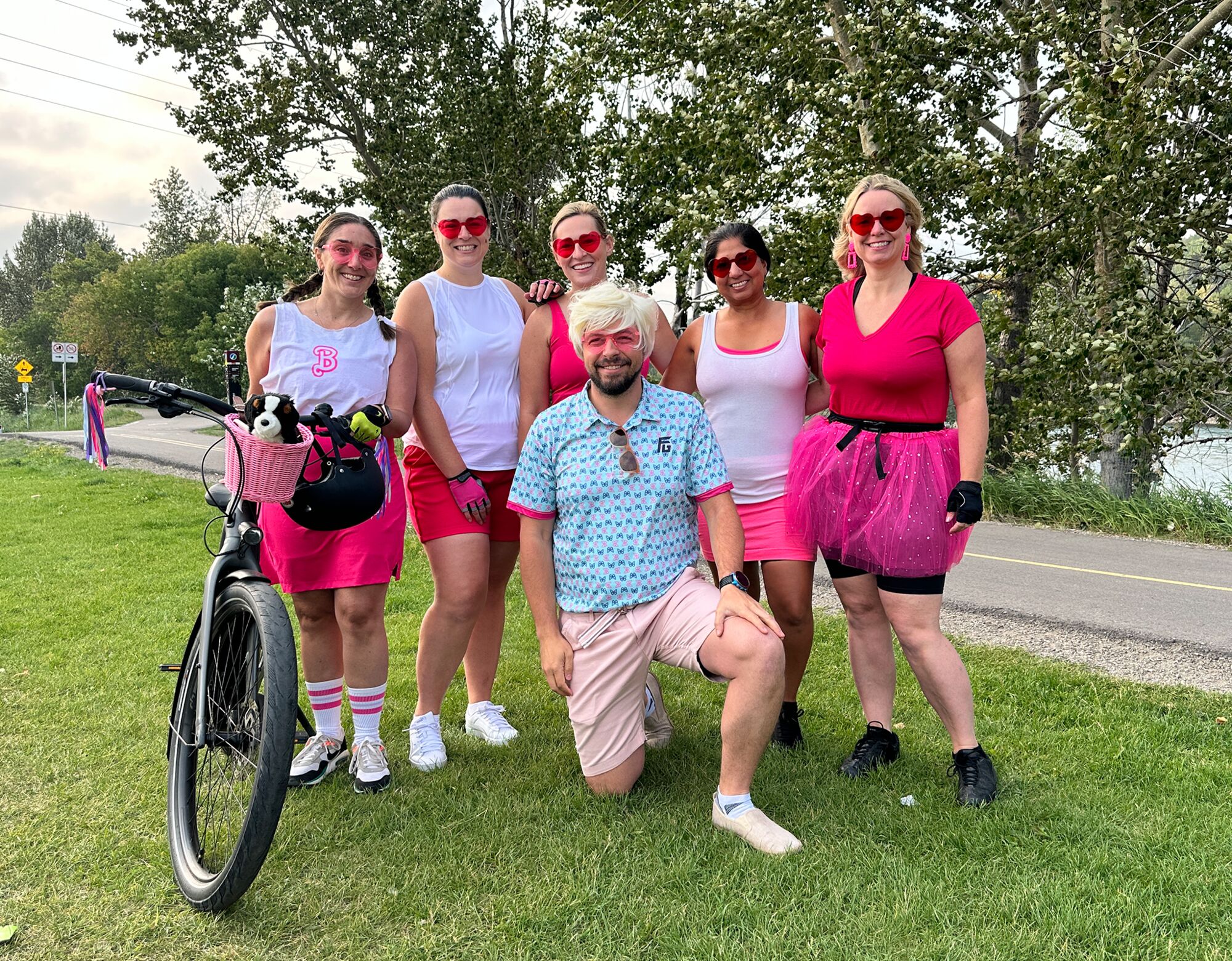 Group of five women and one male standing and smiling wearing pink and white Barbie and Ken costumes in summer outdoor setting with bicycle