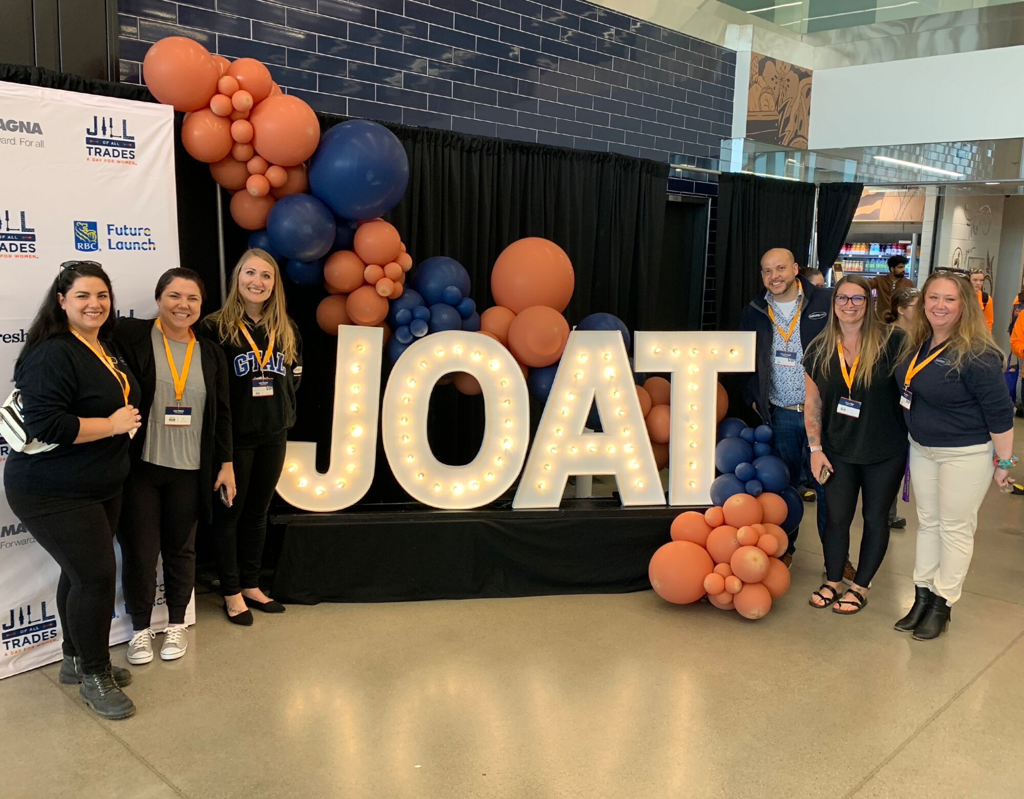 Five women and one male standing beside indoor marquee signage with the letters J.O.A.T smiling at the camera