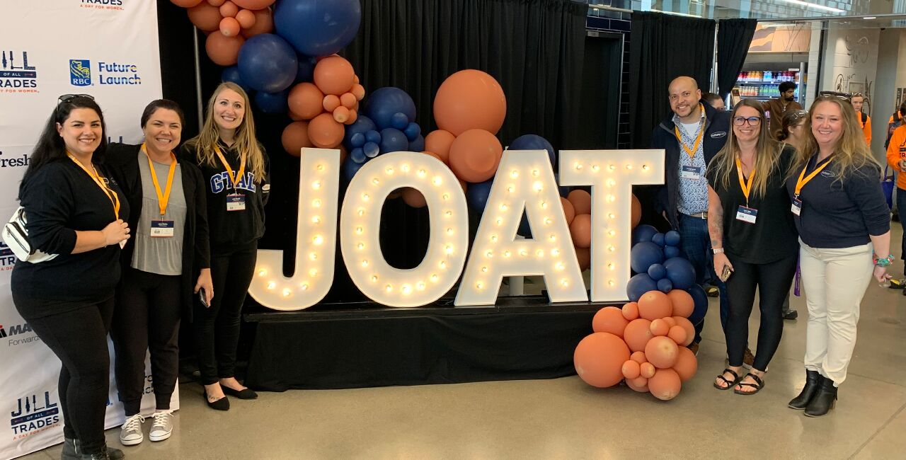 Five women and one male standing beside indoor marquee signage with the letters J.O.A.T smiling at the camera