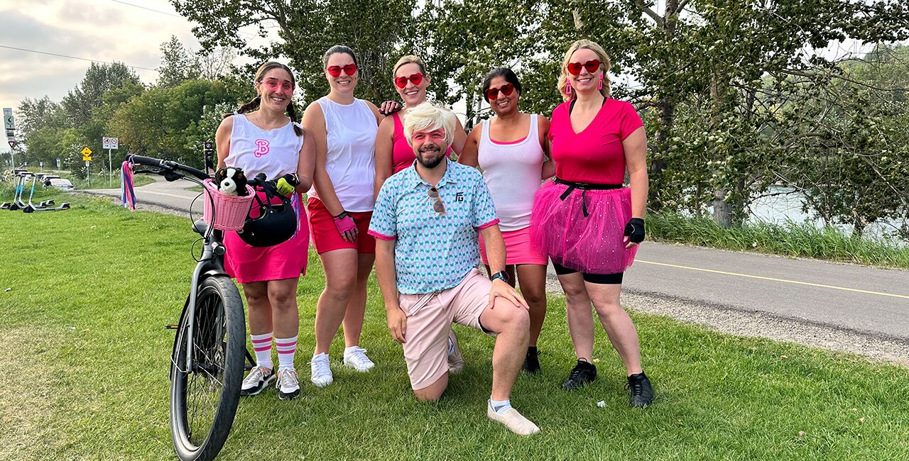 Group of five women and one male standing and smiling wearing pink and white Barbie and Ken costumes in summer outdoor setting with bicycle