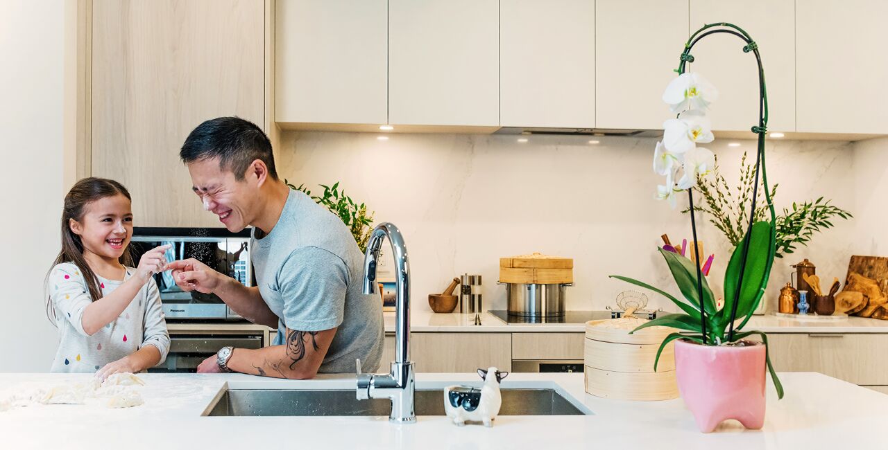 Dad and daughter playing with dough and laughing while leaning against the kitchen island with white oak cabinets, silver faucet and orchid on the counter