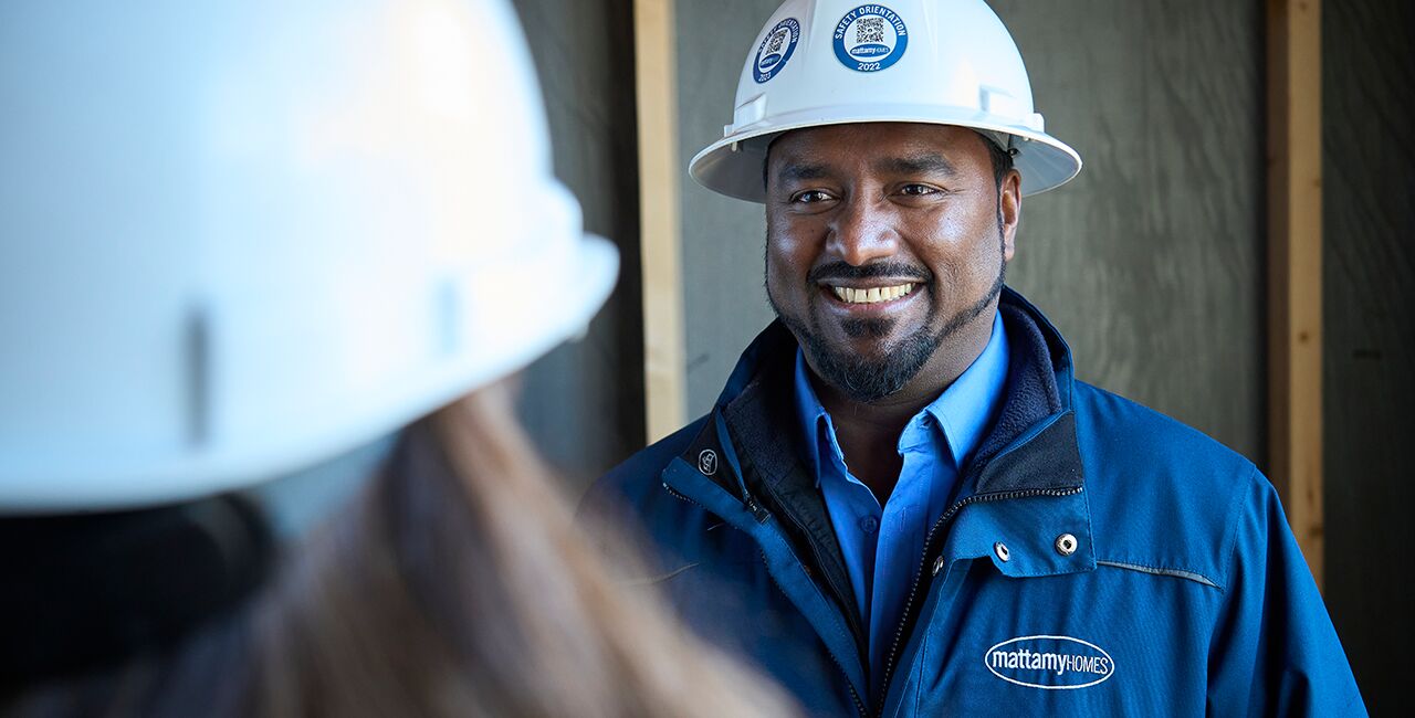 Two builders wearing blue Mattamy jackets and white hard hats have a conversation inside of a framed house.