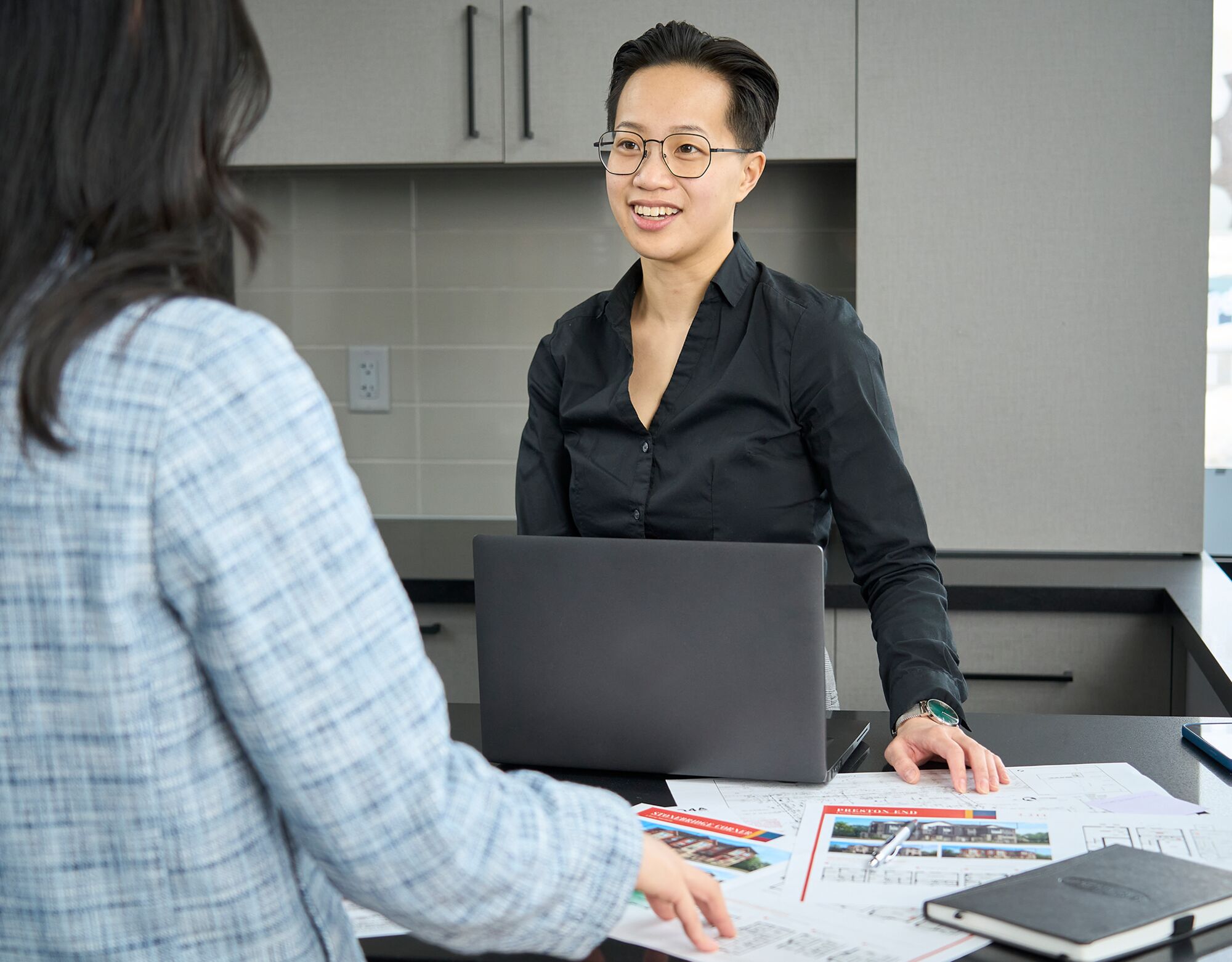 Two women in an office environment having a conversation while reviewing floorplans spread out on a table with an open laptop and a notebook. 