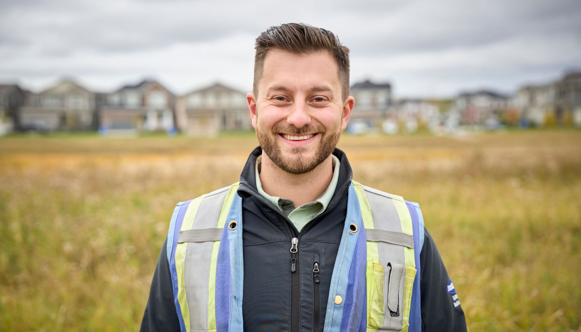 Male employee wearing safety vest smiling at camera while outside with grass and houses blurred behind him